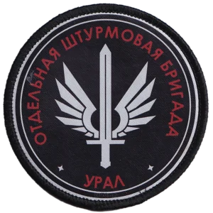 Home 15 Ural Assault Brigade Patch with red Cyrillic unit lettering, silver winged sword emblem, black circular field, and modern Russia–Ukraine conflict context.